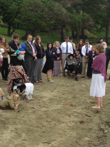 image of a gathering on a beach with an 8 year old in a camping chair and white baptism outfit and her mother in front of her giving a talk