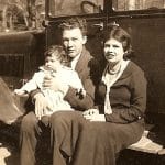 Black and white photograph of man and woman sitting in front of car with a small baby on man's lap.