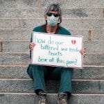 Woman sitting on stairs with sign that reads "how do our battered and tender hearts bear all this pain?"