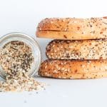 Three slices of everything bagels stacked on top of each other next to a glass jar of everything bagel spices with spice spilling out.