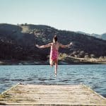 A young girl in a pink swimsuit leaps off a wooden dock into a lake surrounded by hills on a sunny day.