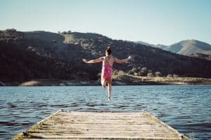 A young girl in a pink swimsuit leaps off a wooden dock into a lake surrounded by hills on a sunny day.