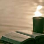 A green cup and open scriptures sit on a wooden beam in front of a body of water with sun shining on it