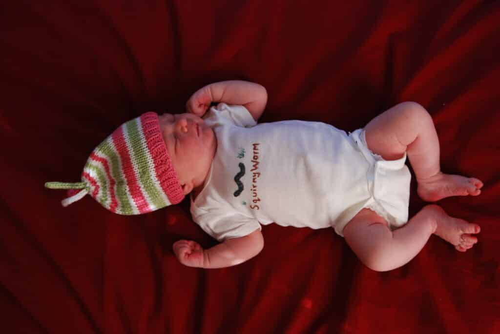 A newborn wearing a home made knit hat and a hand decorated Onesie that says "Squirmy Worm"