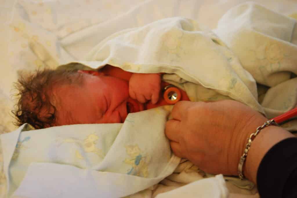 A midwife's hand holds a stethoscope to a crying newborn's chest.