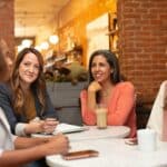 Four women sitting at a cafe table. One is speaking and the other three are listening. Three are women of color.