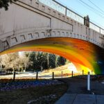 Rainbow painted on the underside of a stone bridge. A pathway goes under the bridge but has a pole barrier to deny access to some