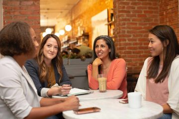 Four women sitting at a cafe table. One is speaking and the other three are listening. Three are women of color.