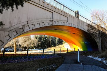 Rainbow painted on the underside of a stone bridge. A pathway goes under the bridge but has a pole barrier to deny access to some